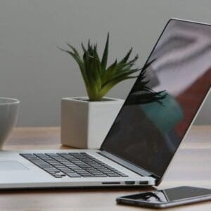 A laptop on a wooden table next to a phone and a plant.